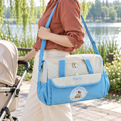 Sac à langer bleu clair à petits pois avec poche latérale froncée, bandoulière bleue et écusson ourson, porté à l'épaule près d'une poussette au bord d'un lac