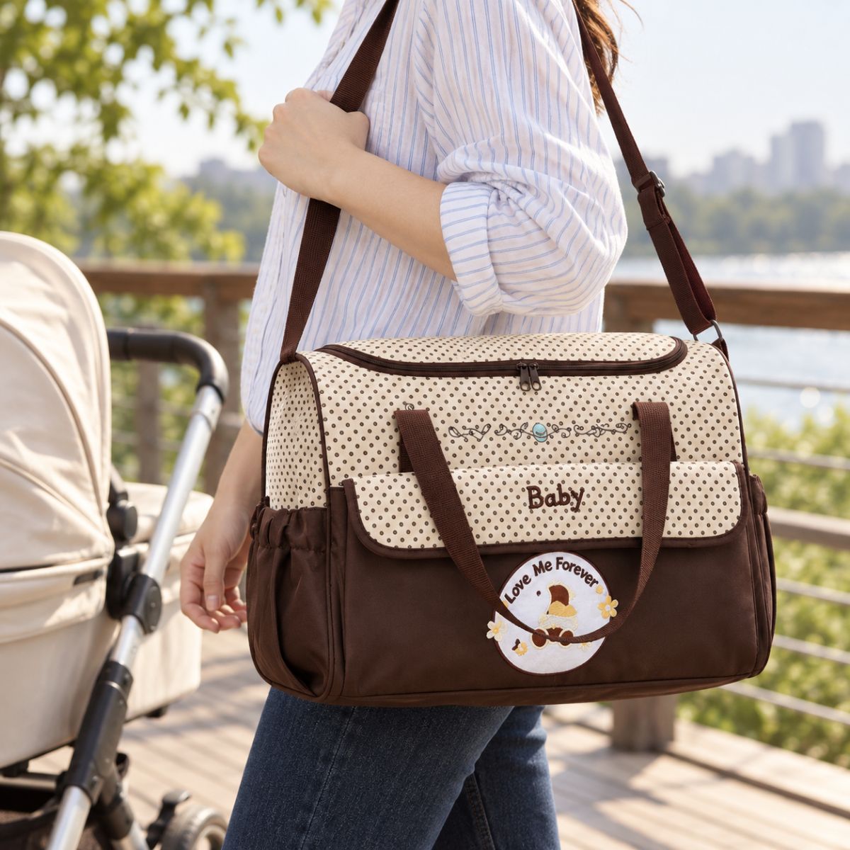 Sac à langer marron à pois avec inscription Baby, poche frontale à rabat et bandoulière, porté à l'épaule près d'une poussette au bord de l'eau