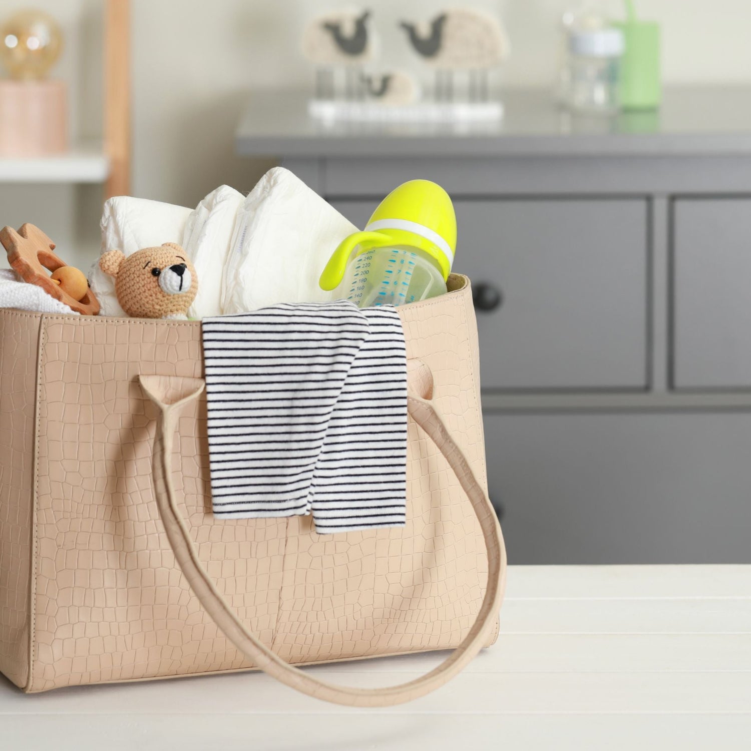 Sac à langer beige rempli d’accessoires pour bébé – couches, biberon, doudou et lange rayé – ambiance douce signée MamanPépite.

