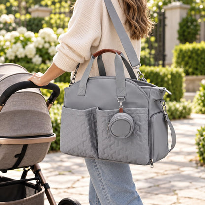 Sac à langer gris clair porté à l’épaule par une maman près d’une poussette, avec pochette ronde et poche latérale zippée visibles.