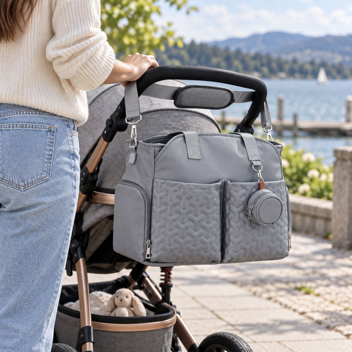 Sac à langer gris clair suspendu à une poussette au bord d’un lac, avec deux poches avant matelassées et étui à tétine.