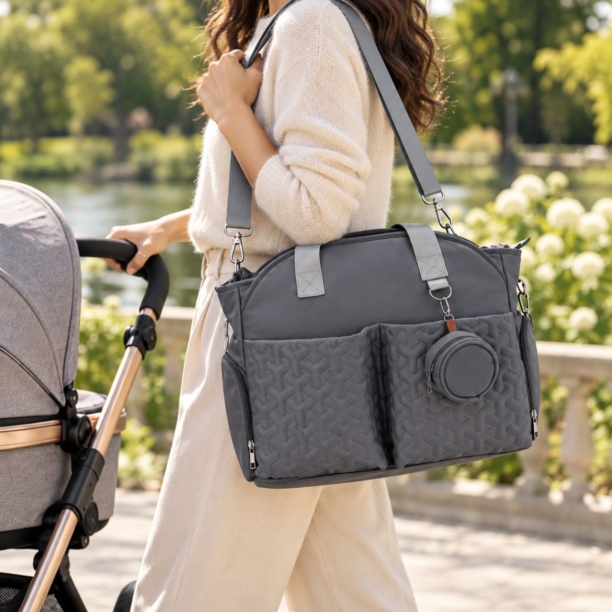Sac à langer graphite porté à l’épaule par une maman au parc, avec deux poches avant matelassées et étui à tétine rond.
