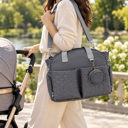 Sac à langer graphite porté à l’épaule par une maman au parc, avec deux poches avant matelassées et étui à tétine rond.