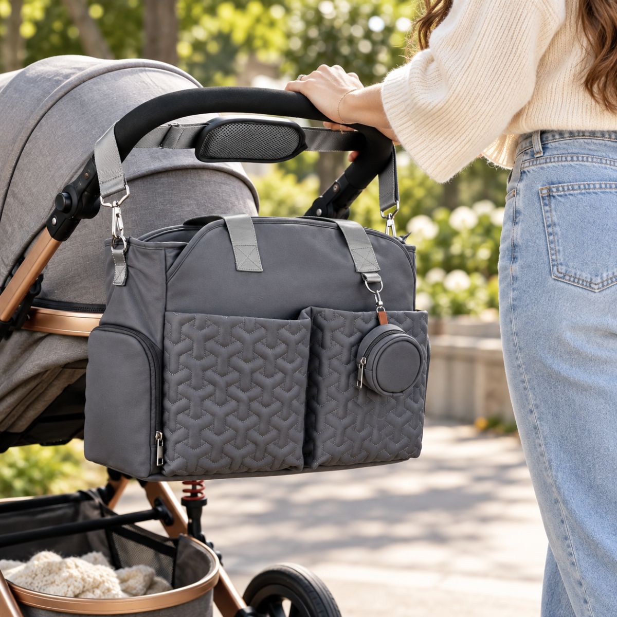 Sac à langer graphite suspendu près d’une poussette grise, avec poches avant matelassées et étui à tétine assorti.