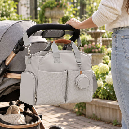 Sac à langer gris perle suspendu près d’une poussette, avec poches avant, fermeture latérale et étui à tétine visibles.