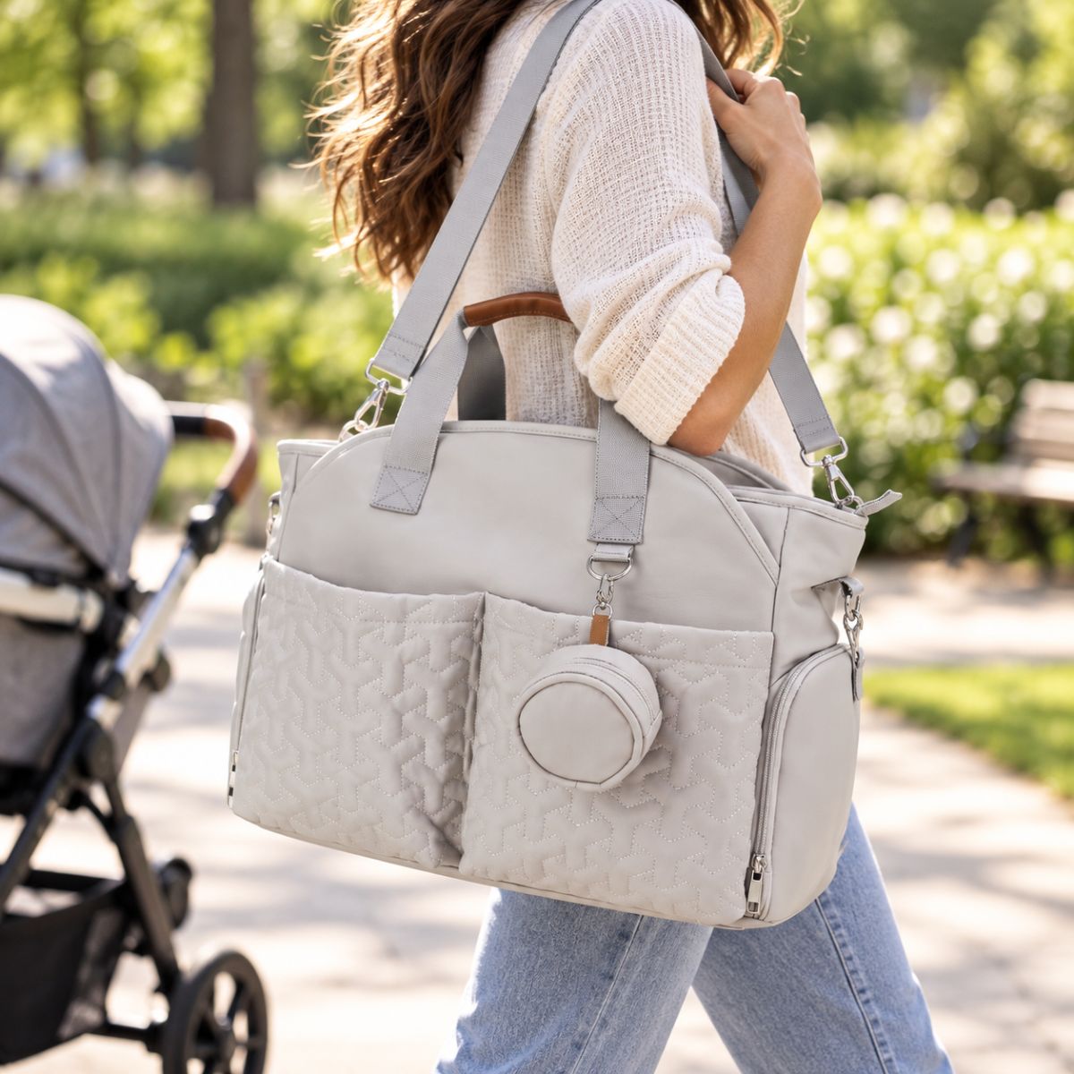 Sac à langer gris perle porté à l’épaule dans un parc, avec deux poches matelassées et étui à tétine rond assorti.
