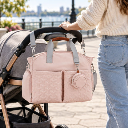Sac à langer rose poudré suspendu près d’une poussette, avec bandoulière grise et étui à tétine assorti.