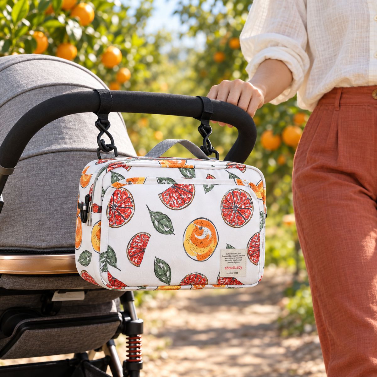 Sac à langer compact blanc à motif d'agrumes rouges et orangés avec feuilles vertes, suspendu au guidon d'une poussette par deux attaches, en extérieur dans un verger