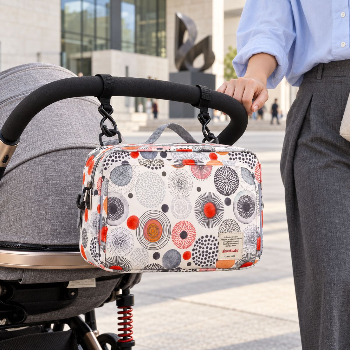 Sac à langer compact blanc cassé à motifs de cercles abstraits noirs, gris, rouges et orange, suspendu au guidon d'une poussette par deux attaches, en extérieur devant un bâtiment moderne