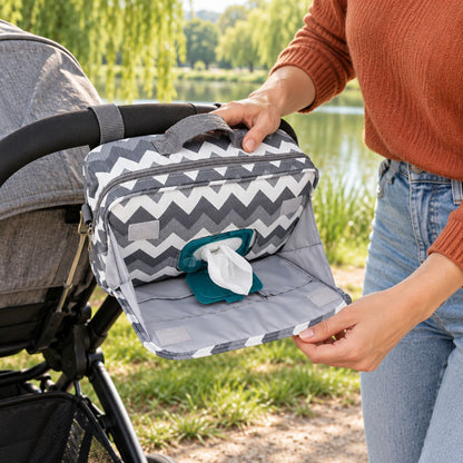 Sac à langer compact à motif chevrons gris et blanc, rabat avant ouvert montrant l'accès aux lingettes et la doublure grise, tenu près d'une poussette en extérieur au bord d'un lac