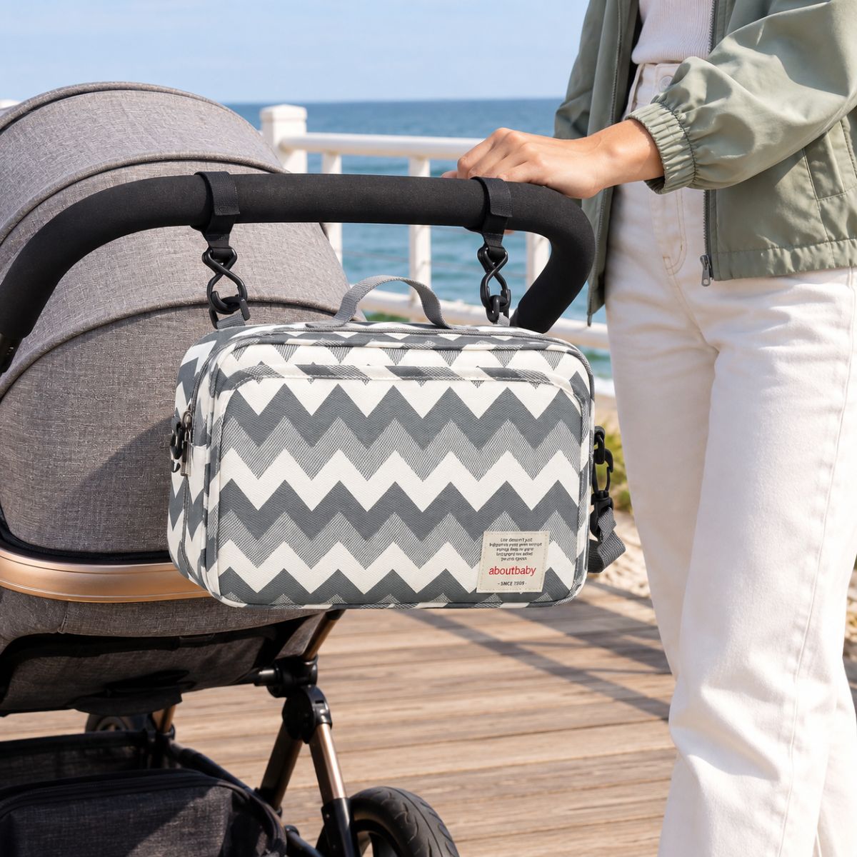 Sac à langer compact à motif chevrons gris et blanc, suspendu au guidon d'une poussette par deux attaches, vue de face en extérieur au bord de la mer