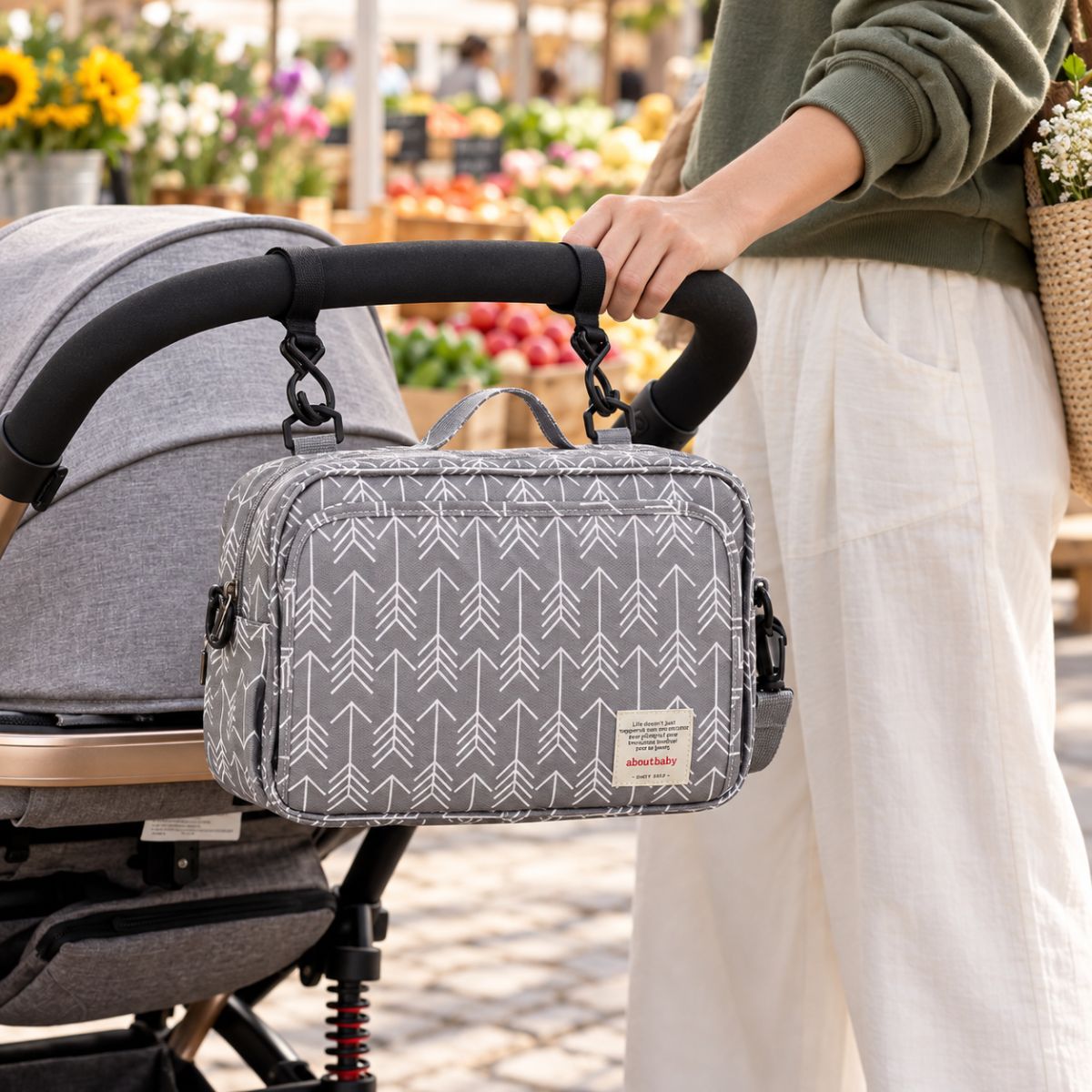 Sac à langer compact gris à motif de flèches blanches, suspendu au guidon d'une poussette par deux attaches, vue de face en extérieur près d'un marché fleuri