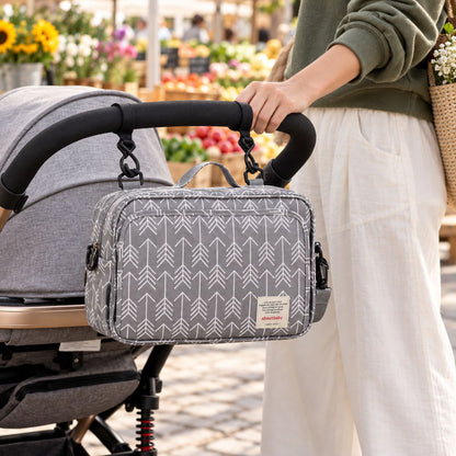 Sac à langer compact gris à motif de flèches blanches, suspendu au guidon d'une poussette par deux attaches, vue de face en extérieur près d'un marché fleuri