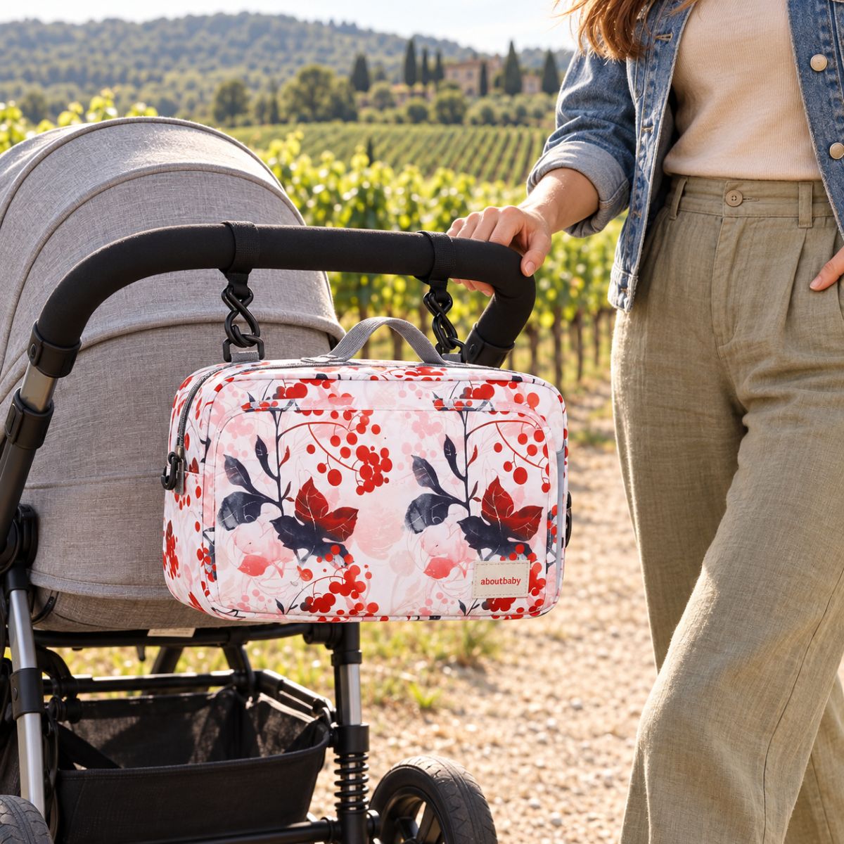 Sac à langer compact à motif floral rose pâle, baies rouges et feuilles foncées, suspendu au guidon d'une poussette par deux attaches, vue de face en extérieur près des vignes