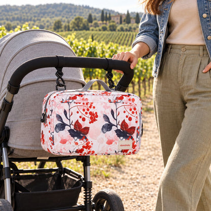 Sac à langer compact à motif floral rose pâle, baies rouges et feuilles foncées, suspendu au guidon d'une poussette par deux attaches, vue de face en extérieur près des vignes