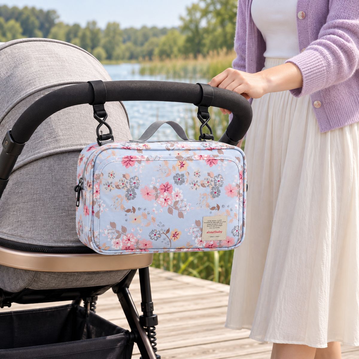 Sac à langer compact bleu ciel à motifs de fleurs roses, blanches et grises, suspendu au guidon d'une poussette par deux attaches, vue de face en extérieur au bord d'un lac