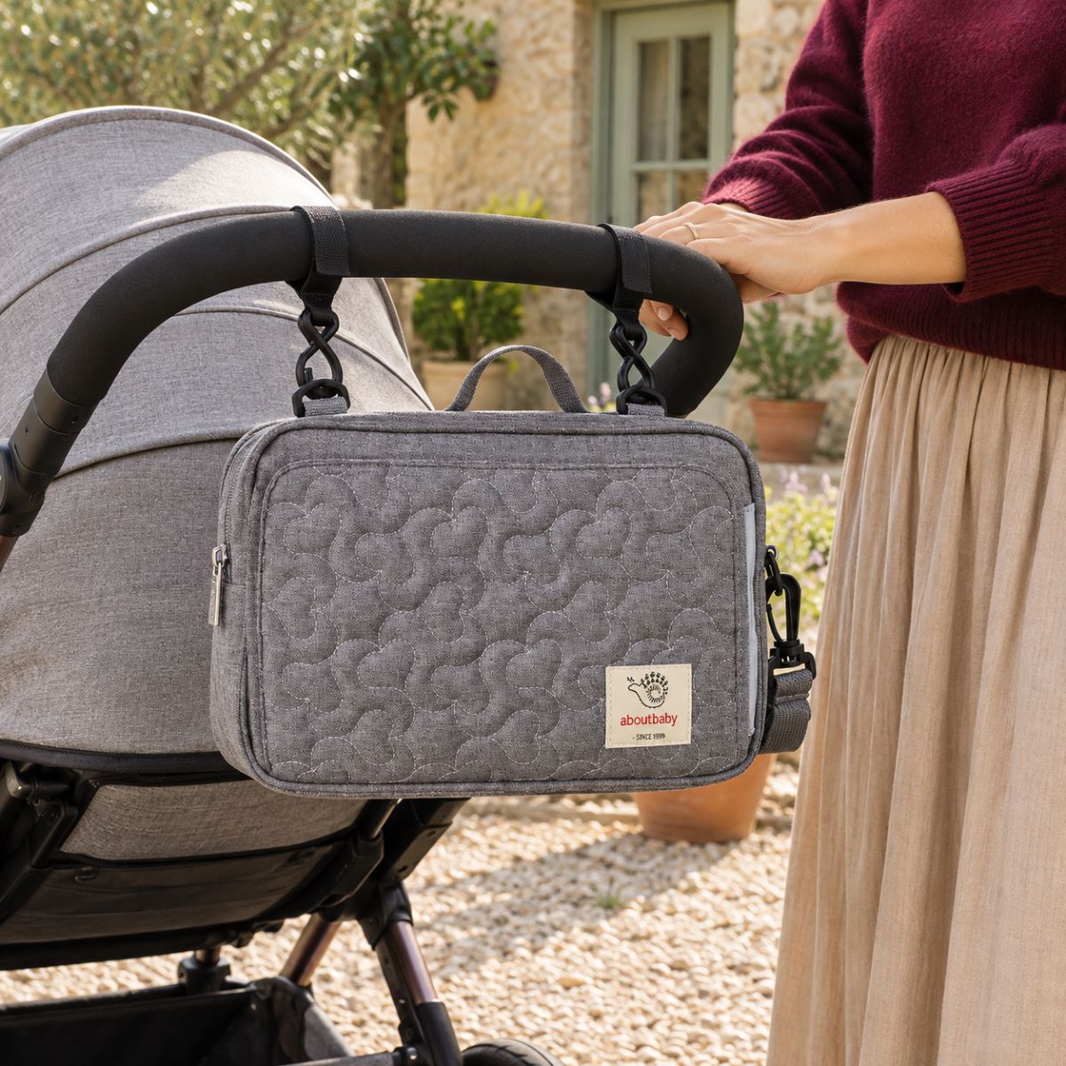Sac à langer compact gris matelassé à motifs floraux ton sur ton, suspendu au guidon d'une poussette par deux attaches, vue de face en extérieur