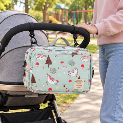 Sac à langer compact vert menthe à motifs de licornes roses et blanches, arcs-en-ciel et sapins, suspendu au guidon d'une poussette par deux attaches, dans un parc avec aire de jeux