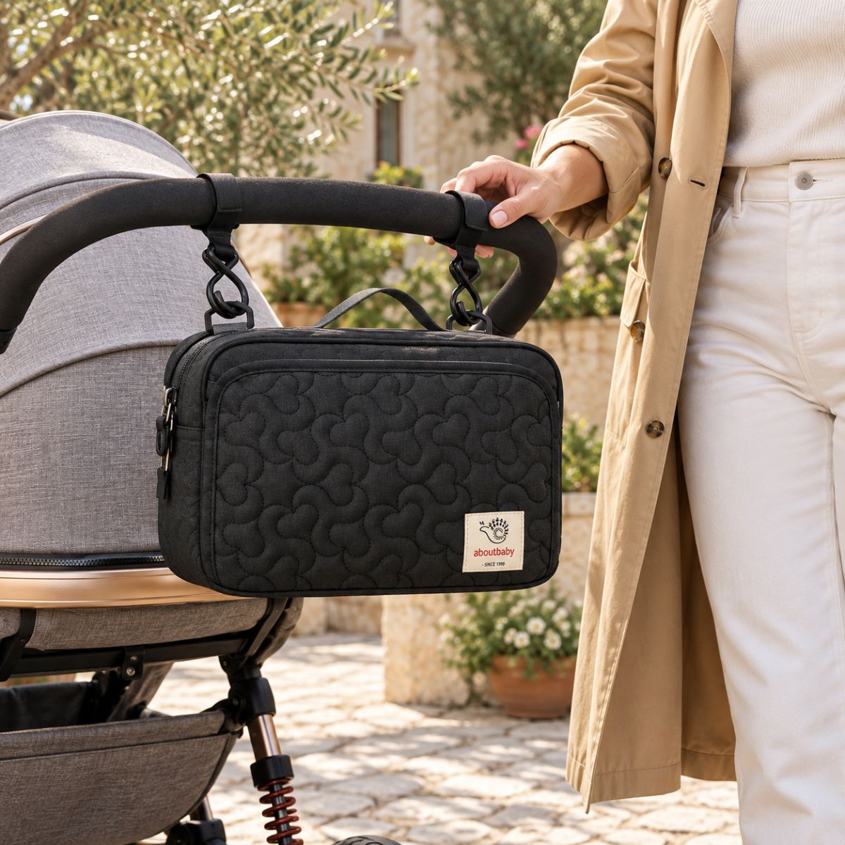 Sac à langer compact noir matelassé à motifs floraux ton sur ton, suspendu au guidon d'une poussette par deux attaches, vue de face en extérieur