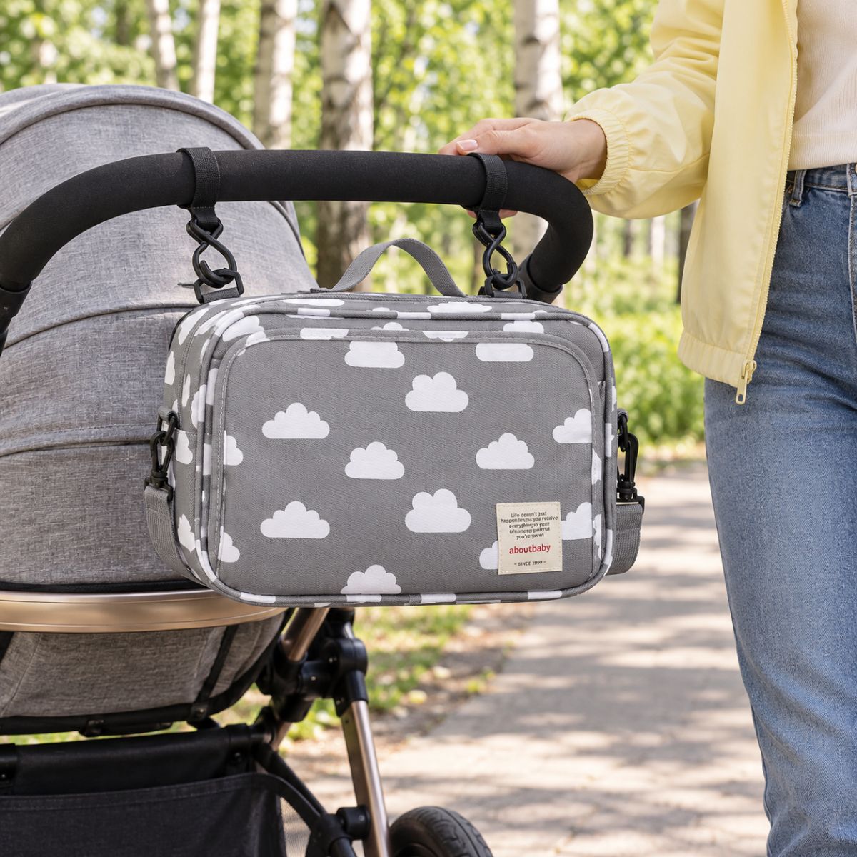 Sac à langer compact gris à motif de nuages blancs, suspendu au guidon d'une poussette par deux attaches, vue de face en extérieur dans un parc