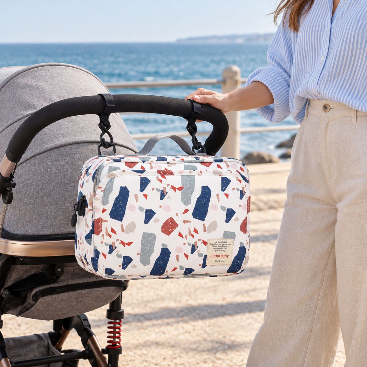 Sac à langer compact à motif terrazzo multicolore sur fond blanc cassé, suspendu au guidon d'une poussette par deux attaches, en bord de mer
