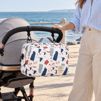 Sac à langer compact à motif terrazzo multicolore sur fond blanc cassé, suspendu au guidon d'une poussette par deux attaches, en bord de mer