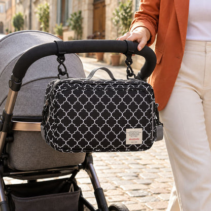 Sac à langer compact noir à motif de treillis géométrique blanc, suspendu au guidon d'une poussette par deux attaches, vue de face en extérieur dans une rue pavée