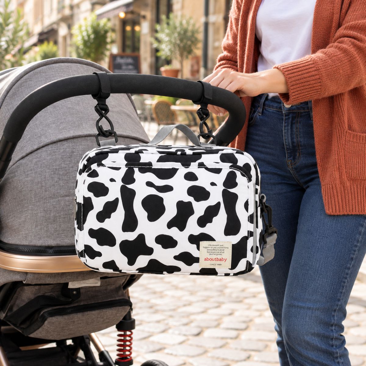 Sac à langer compact à motif vache noir et blanc, suspendu au guidon d'une poussette par deux attaches, vue de face en extérieur