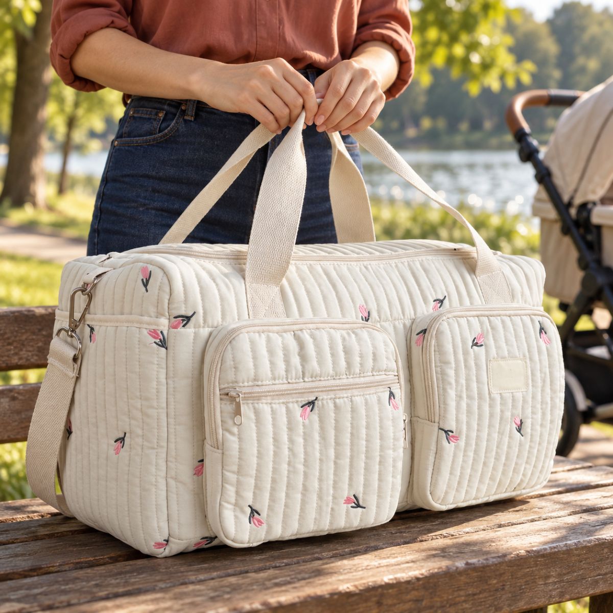 Sac à langer en tissu crème matelassé à petites tulipes roses brodées, deux poches avant zippées et bandoulière beige, présenté sur une table en bois près d'une poussette au bord d'un lac