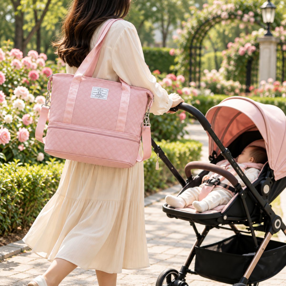 Sac à langer extensible rose porté à l’épaule par une femme près d’une poussette dans un parc fleuri