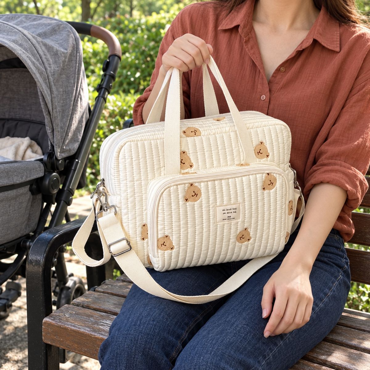 Sac à langer ourson brodé en tissu crème matelassé, avec poche avant zippée et bandoulière réglable, posé sur les genoux