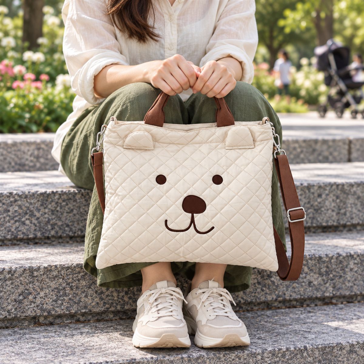 Vue de face d’un sac à langer fourre-tout beige matelassé, décoré d’un visage d’ours et de petites oreilles en relief, tenu par ses anses dans un parc