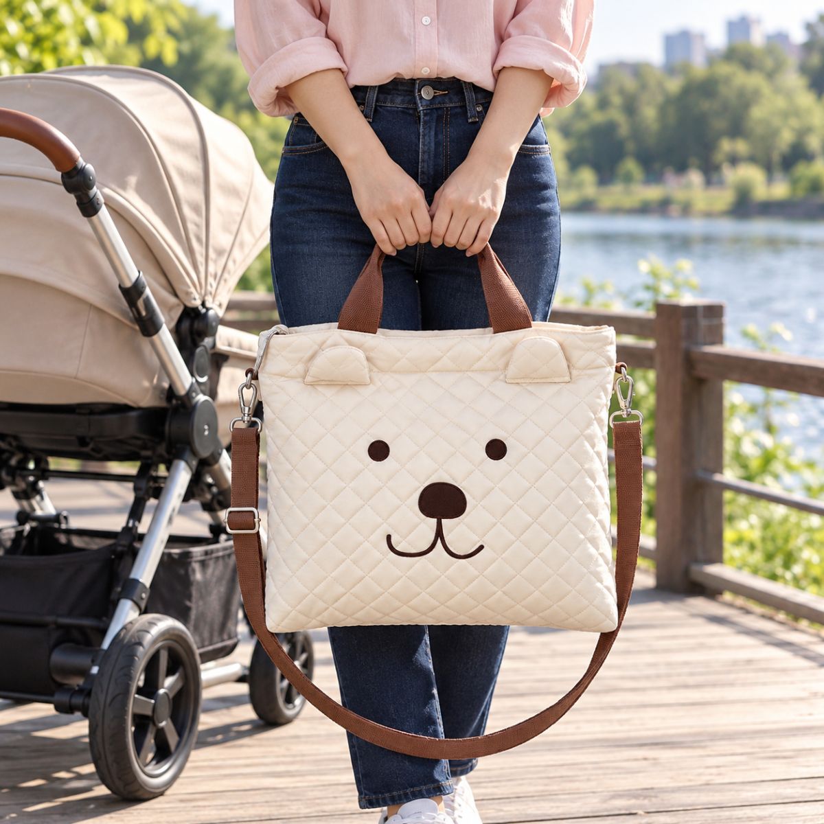 Sac à langer fourre-tout beige crème à motif ours, porté à la main par ses anses marron, avec bandoulière visible près d’une poussette au bord d’un lac