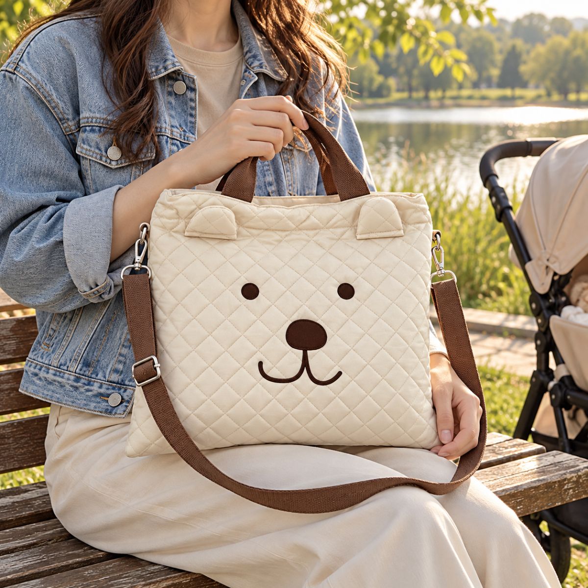 Sac à langer beige à visage d’ours marron, tenu par ses deux anses sur un banc au bord d’un lac, avec bandoulière amovible et poussette à proximité