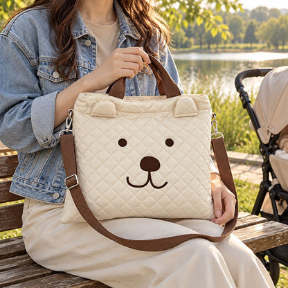 Sac à langer beige à visage d’ours marron, tenu par ses deux anses sur un banc au bord d’un lac, avec bandoulière amovible et poussette à proximité