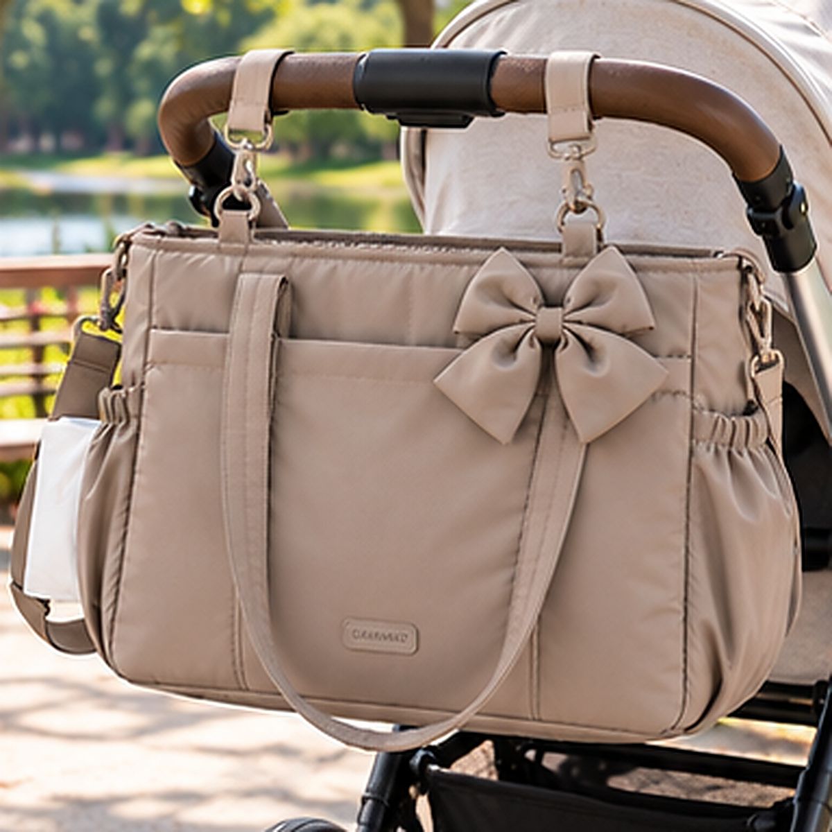 Sac à langer camel avec nœud décoratif, anses et poches latérales, présenté sur une poussette beige au parc