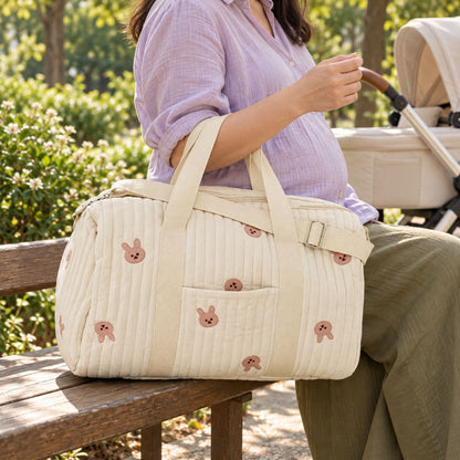 Sac à langer polochon crème à petits lapins roses, avec poche extérieure plaquée et anses claires, tenu par une femme assise sur un banc près d'une poussette