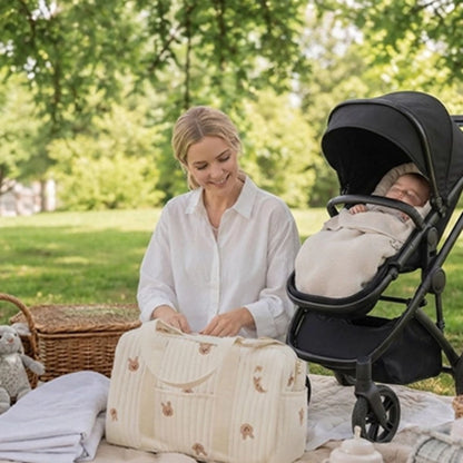 Sac à langer polochon crème à motifs lapins roses, ouvert par une maman installée avec son bébé sur une couverture de pique-nique près d'une poussette