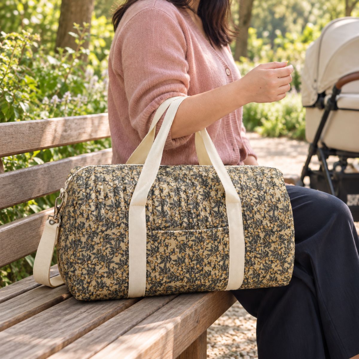 Sac à langer polochon matelassé à imprimé botanique de feuilles et petits fruits jaunes, porté par une femme assise sur un banc dans un parc