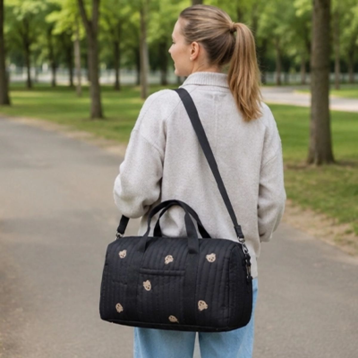 Sac à langer polochon noir à motifs oursons, porté en bandoulière par une femme de dos pendant une promenade dans un parc