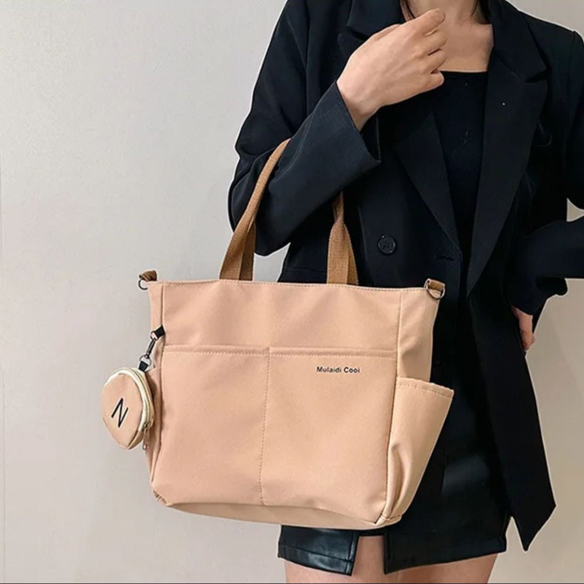 Sac a langer kaki porté à l’épaule par une maman en veste noire, pochette ronde attachée